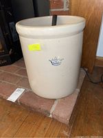 Off-white 6 gallon stoneware crock with blue crown mark visible, placed on brick hearth near wood floor and wall.