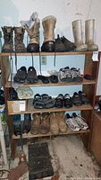Photo shows a 4-tier wooden shelf holding various men's footwear including boots, sandals, sneakers and a pair of gloves. Mostly men's size 8 or 9.