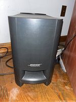 Front view of black Bose subwoofer with logo and vented opening on wooden floor, showing physical condition.