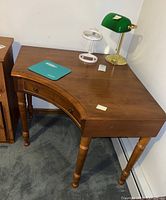 Corner wooden desk with curved front, turned legs, small drawer, green desk lamp, white magnifier stand, and blue mouse pad on top.