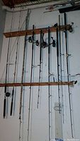Eight fishing poles hung on two wooden racks mounted on a wall, showing various types and reels attached, condition weathered.