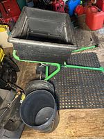 Side view of the John Deere fertilizer spreader showing black plastic hopper, green metal frame, and two wheels on rubber tires. Includes a bucket placed next to it for size reference.