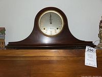 Full view of the mantel clock resting on a wooden surface showing its arched dark wood case and round face.