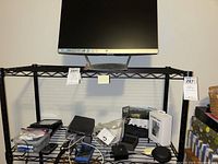 HP Pavilion monitor on black metal shelf with multiple electronic items below it including phone accessories, GPS unit box, and cables.