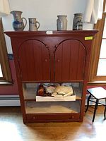 Front view of vintage red painted wood cabinet with closed double arched doors, open lower shelf space with box containing household items, and bottom drawer closed. Stoneware jugs are on top of the cabinet.