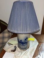 Full view of the pottery lamp with pleated blue lampshade on wooden surface with cloth underneath.