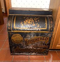 Front view of vintage black metal storage bin with orange and yellow floral design and B. Fischer & Co. branding, showing wear and scratches.
