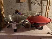 Two electric woks side by side: one stainless steel with glass lid and one red with matching lid, both on black electric heating bases. A clear glass bowl and white ceramic mixing bowls visible behind the woks.