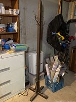 Full view of tall wooden coat rack standing upright in basement, showing vertical post and base feet, surrounded by basement items.