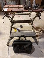 Full view of Black & Decker Workmate workbench with wooden top showing three metal hand planes and a black tool bag below it on the floor in a basement setting.