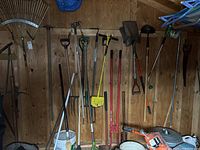 Wide shot of assorted yard tools hanging on wooden wall inside shed, including rakes, spades, pitch forks, and hole digger.