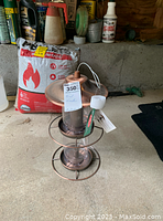 Photo of a new copper bird feeder with circular perch and hanging wire, sitting on a concrete floor with gardening and household items in the background.
