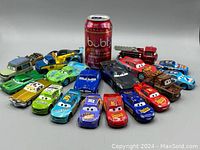 Front view of 17 toy cars arranged with beverage can for scale, displaying various character cars from Cars movie with colors red, blue, green, gold, brown, black.