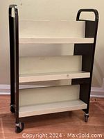 Front angle view of large double-sided metal book cart with cream shelves and brown frame on caster wheels, showing three sloped shelves on one side.