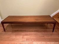 Full front view of rectangular walnut coffee table on hardwood floor against beige wall, showing table legs and overall structure.