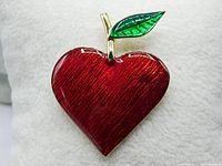 Close-up front view of the red heart/apple brooch showing the enamel texture and green leaf on gold metal.