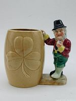 Side view of ceramic mug with beige color and embossed clover leaf design on the cup, showing leprechaun figurine handle in bright colors.