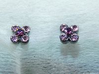 Pair of amethyst-colored crystal flower stud earrings lying flat on a white surface, showing their faceted crystals and flower-shaped design.