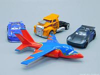 Four toy vehicles displayed including a yellow and purple Hot Wheels semi truck, two Disney Pixar Cars plastic cars, and a blue and red Black Falcon Hawk plane from Cars Toons.