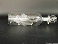 Side view of clear glass bottle with hand blown sailing ship inside and glass stopper on black surface.