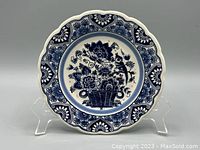 Front view of the vintage Delft Blue ceramic plate showing blue floral and bird designs with a scalloped edge on a clear display stand.