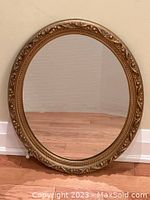 Full view of the vintage gold oval mirror, showing the shape, gold ornate frame, and mirror surface on hardwood floor background.