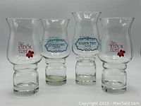 Four clear hurricane glasses side-by-side, two with red logos and two with blue logos, showing slight height variation