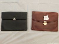 Two leather portfolio cases, black and brown, flat on a surface showing front flap side with metal clasps.