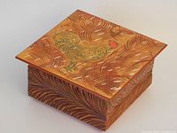 Top angled view of square handmade wooden box with painted rooster on lid and carved textured sides.