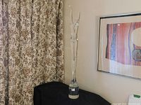 Full view of the tall clear crystal sculpture next to a patterned curtain and wall art.