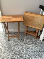 Two wooden folding TV tables shown side by side, one set up with legs extended, another folded upright against the wall. Light wood with parquet pattern tops.