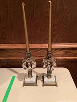 Pair of candlesticks with marble bases, brass stems decorated with clear crystal pendants, each holding a gold candle on top, photographed on a flat surface with wood panel background.