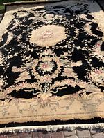 Full view of black wool rug with cream border and central floral medallion laid flat outdoors