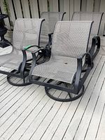 Four black metal frame outdoor swivel chairs with woven fabric seats arranged on a wooden deck against a fence.