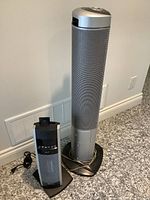 Both standing tower appliances shown side by side on floor, one taller and wider with attached remote on back, other smaller with buttons visible on front.