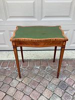 Front and angled view of vintage wooden accent table with green leather top showing wear, highlighting curved edges and wood inlay.