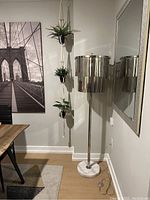 Full view of the floor lamp positioned by a wall, showing the solid round white marble base, polished metal pole, and two-tier chandelier shade made of vertical metal segments.