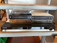 Complete lot showing 4 boxed Game of Thrones DVD sets stacked on a Samsung DVD player unit, showing front of player and DVD set spines.