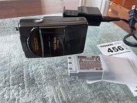 Konica digital camera standing on its side showing front and side with one DR-LB3 battery in foreground and charger in background.