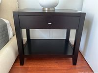 Front view of modern espresso finish solid wood end table showing drawer and lower shelf, next to beige upholstered furniture on a wood floor.