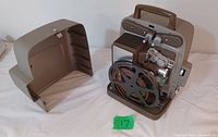 Photo of vintage Bell and Howell film projector with protective cover removed, showing metal film reel, manual controls, and carrying handle.