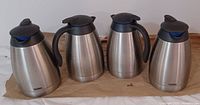 Four stainless steel Thermos coffee serving carafes arranged side by side, showing front and side views with black plastic handles and lids