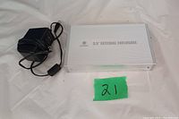 Silver 3.5 inch external USB hard drive enclosure alongside black power adapter for the enclosure. The enclosure is labeled with "3.5\" EXTERNAL ENCLOSURE" and the brand "OCOOLM". The power adapter has a standard plug and cable for providing external power to the enclosure.