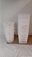 Photo showing seven square white translucent plastic pails with red measurement markings stacked on the right side, and five round similar white plastic pails stacked on the left side.