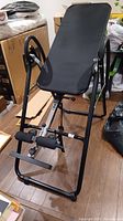 Black inversion table with metal frame, padded backrest, and foam ankle supports on wooden floor in storage area.