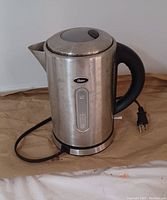 Single image showing a stainless steel Oster electric kettle with black handle and lid knob, connected to a power cord with a plug.