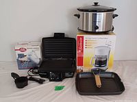 Photo of all kitchen appliances in the lot arranged against a plain background, including boxed coffeemaker, boxed hand mixer, open sandwich grill, grill pan, and measuring cups.