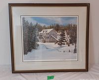 Framed and matted winter scene print showing a watermill house in snow with pine trees and blue sky.