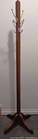 Full view of the tall oak coat rack showing the upright wooden pole with metal hooks and the base standing on the floor.