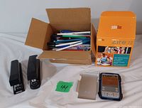 Palm One Zire 72 Palm Pilot, open box of assorted pens, and two black staplers on white cloth background